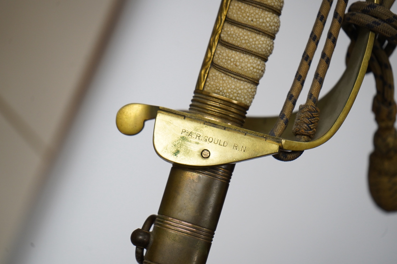 A George VI naval officer’s dress sword by Wilkinson (unnumbered), with regulation brass hilt with folding guard, engraved P.A.R. Gould R.N., bullion dress knot, in its brass mounted leather scabbard and contained in its